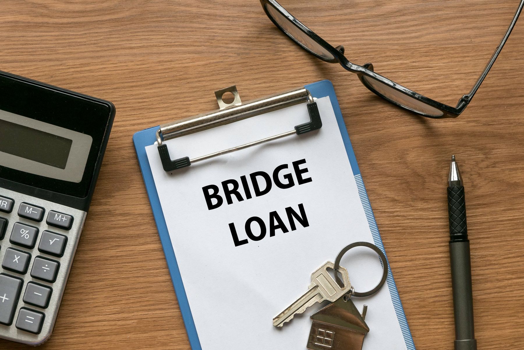 A clipboard marked Bridge Loan on a wooden desk with a calculator, glasses, a pen and a house key